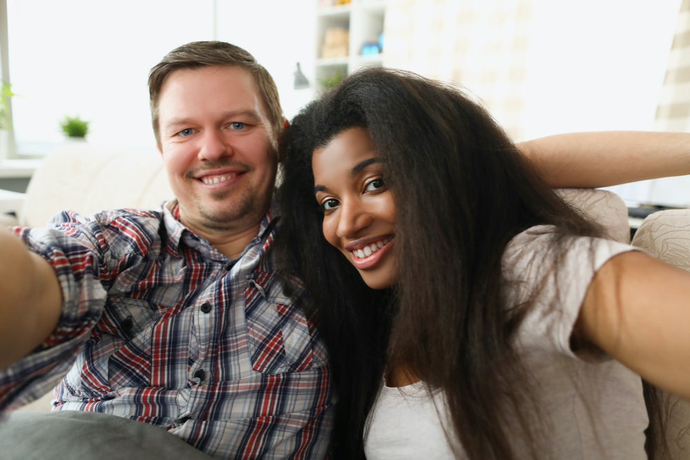 Cheerful multiethnic couple in love sits in embrace in cozy living room at home