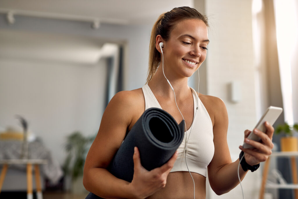 Happy female athlete holding exercise mat while texting on mobile phone at home