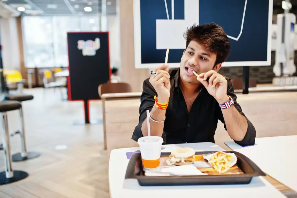 Funny and stylish indian man sitting at fast food cafe and eating french fries show his tongue