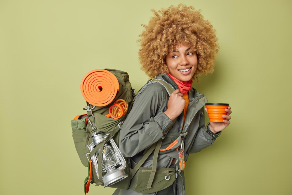 shot of cheerful female hiker dressed in jacket carries rucksack with necessary equipment