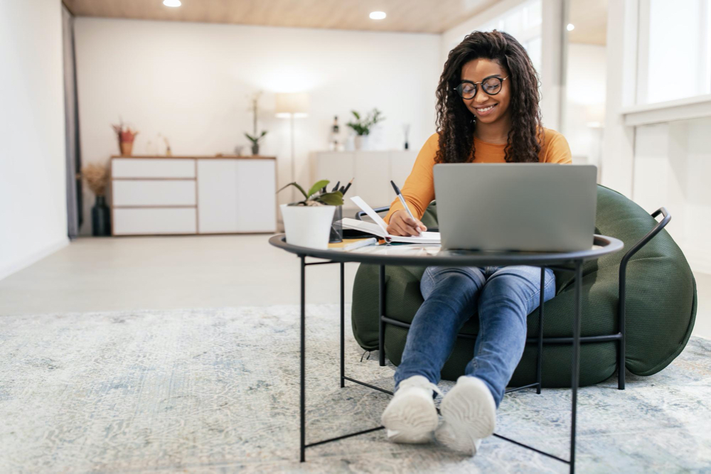 Photo smiling woman sitting at table using computer writing in notebook