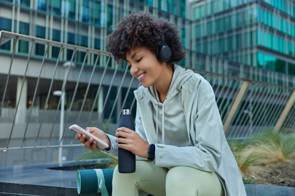 photo woman listens music in headphones scrolls social networks via smartphone takes break after workout poses 