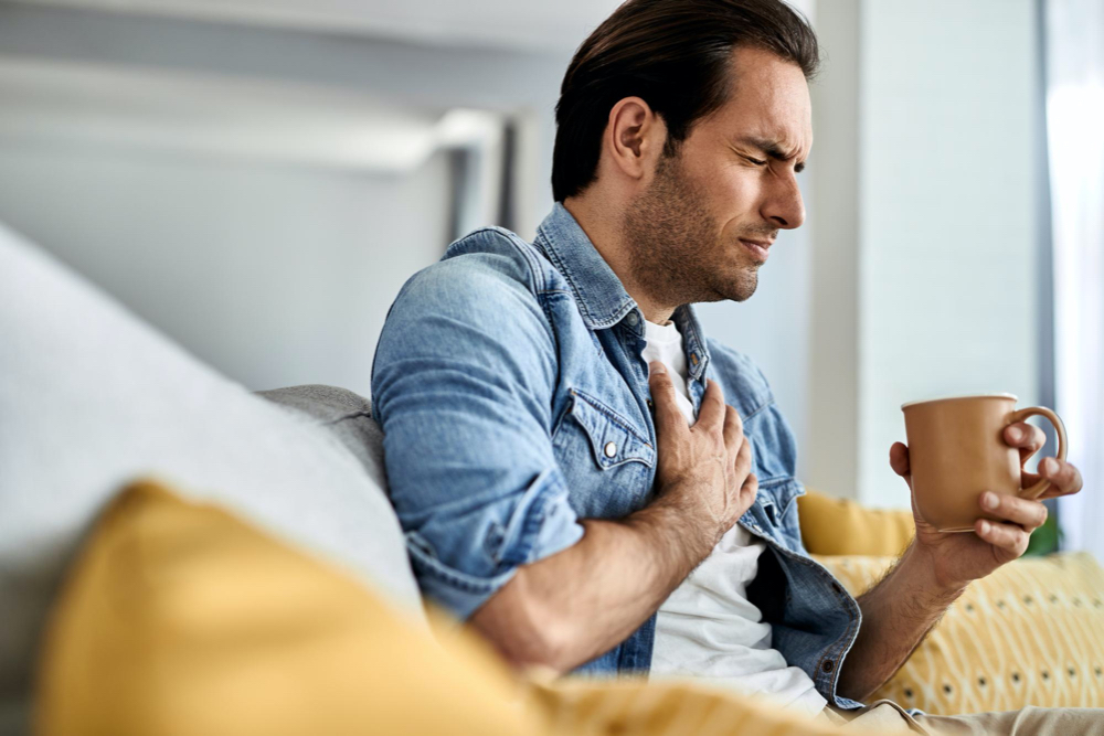 photo young man feeling sick and holding his chest in pain while drinking tea in the living room