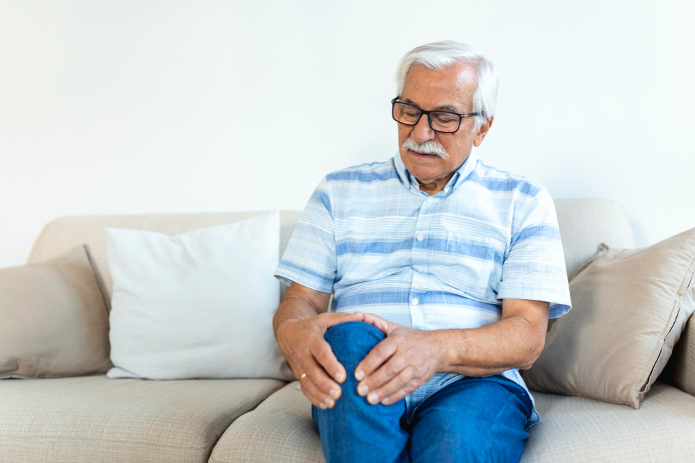 photo elderly man sitting on a sofa at home and touching his painful knee people health care and problem concept unhappy senior man suffering from knee ache at home
