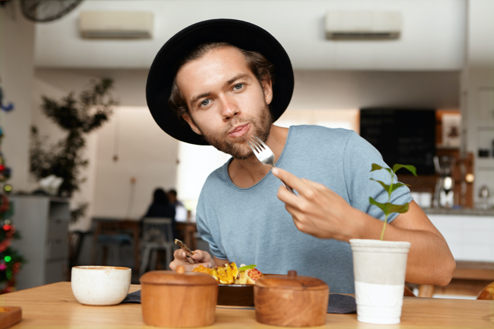 photo people, food and lifestyle concept. indoor shot of attractive young student wearing black hat and blue t-shirt appeasing his hunger
