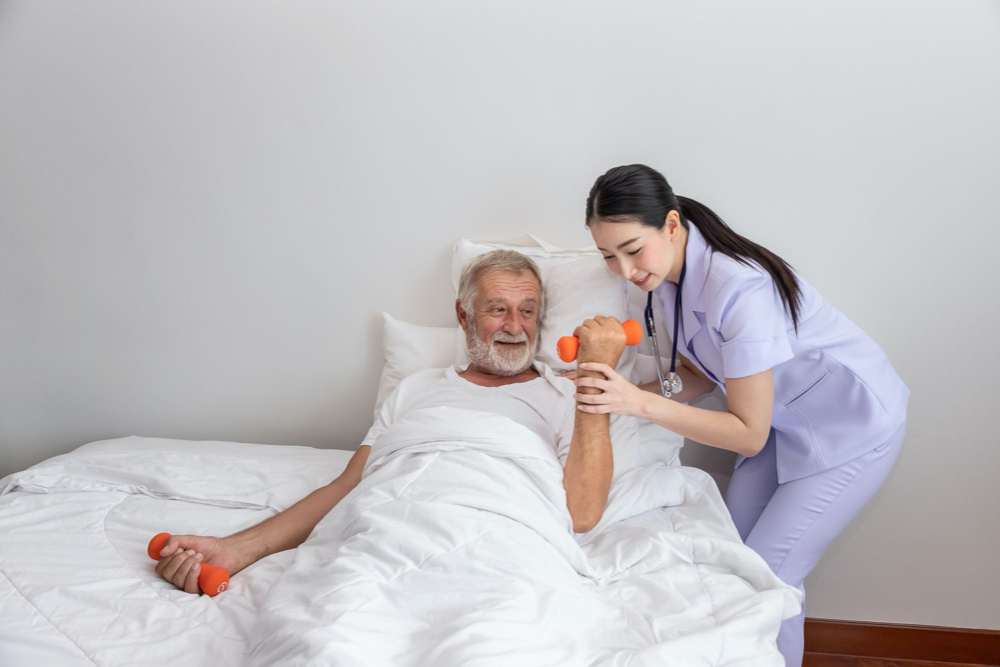 Senior man holding dumbbells with physiotherapist in bedroom at nursing home