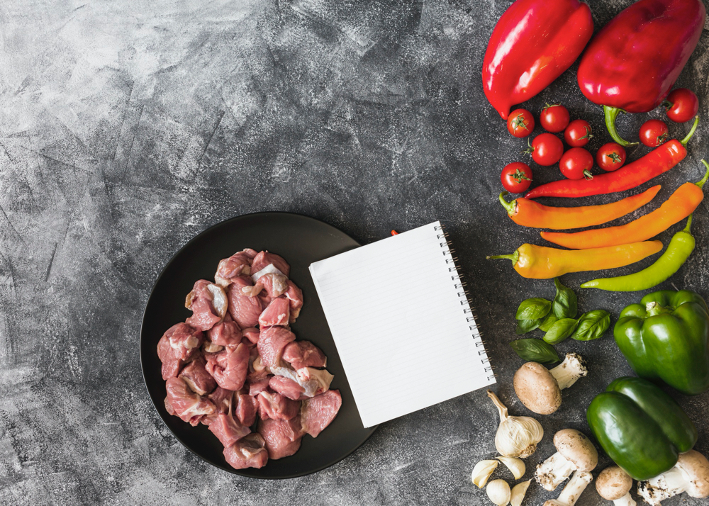 An overhead view of raw meat with notebook and colorful vegetables on stained background