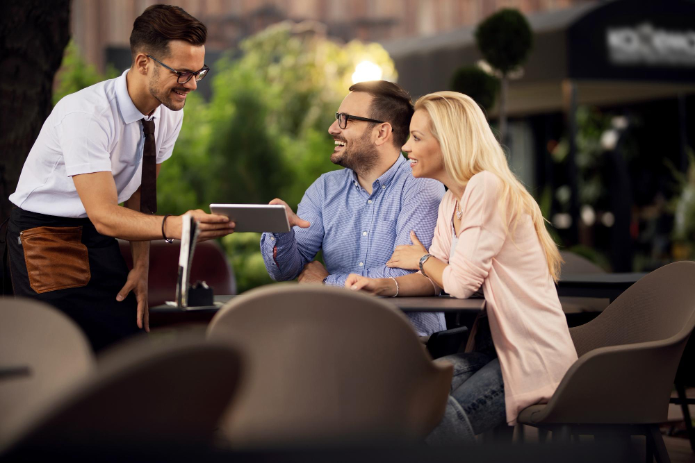 Happy couple making an order in a cafe while waiter is showing them menu on digital table
