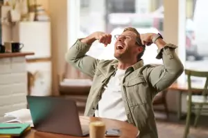 Portrait of pleased smiling young man shouting with rejoice sitting in cafe in front of laptop