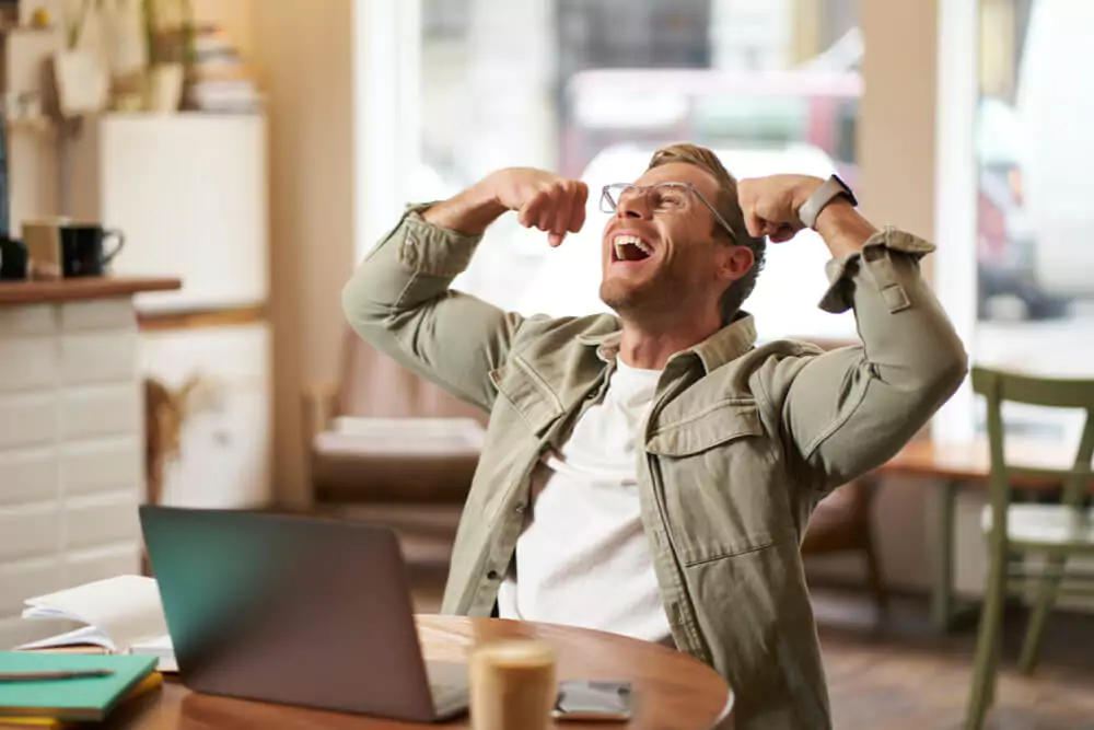 Portrait of pleased smiling young man shouting with rejoice sitting in cafe in front of laptop