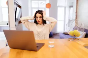 Surprised Pretty young woman is sitting on the kitchen and working on her laptop and mobile phone